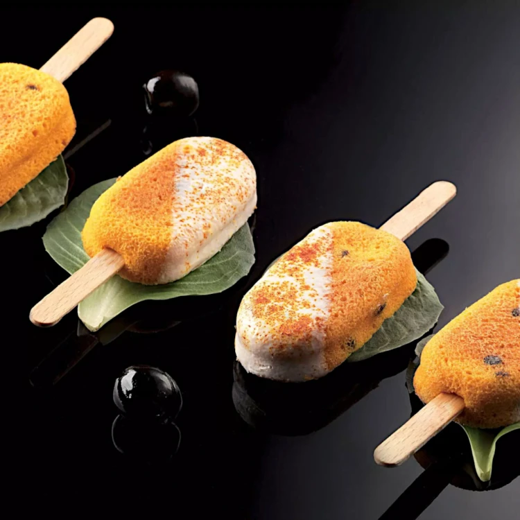 Lody na patyku o smaku mango w dwukolorowej polewie, ułożone na liściach na czarnym tle.