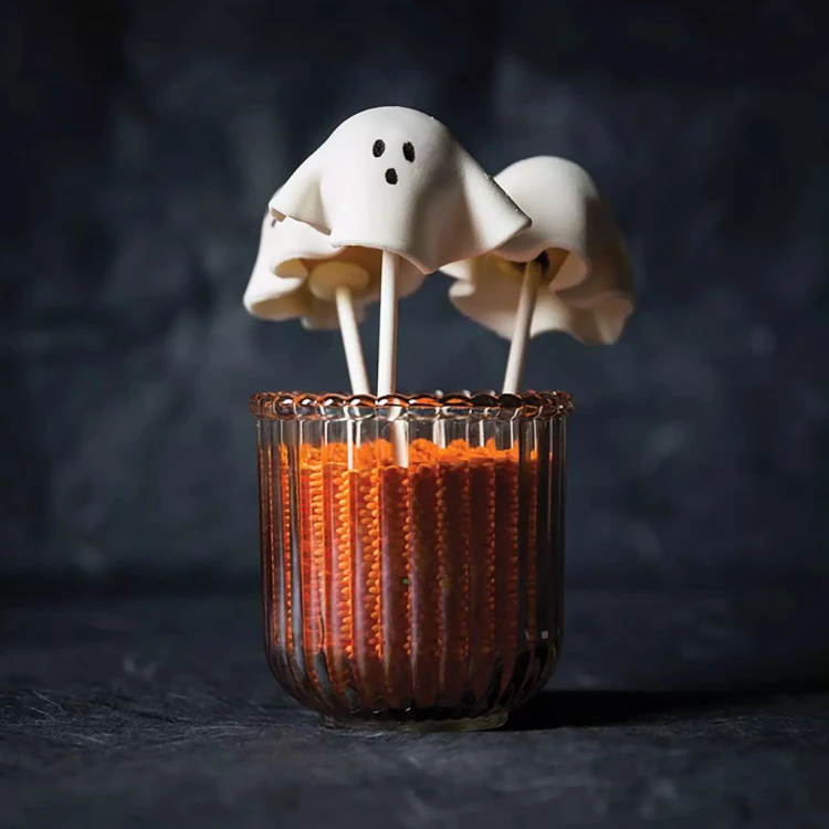 Halloweenowy cakepops w kształcie duszka, ustawiony w szklance z pomarańczową posypką.