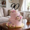 Dwupiętrowy różowy tort ozdobiony pastelowymi kwiatami i czarnym topperem w kształcie liczby 90, ustawiony w eleganckiej aranżacji salonu.