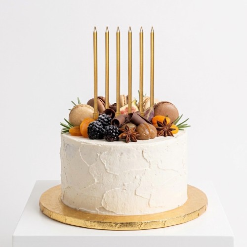Tort z białym kremem ozdobiony owocami i makaronikami, na którego wierzchu ustawiono sześć smukłych złotych świeczek w eleganckiej linii.